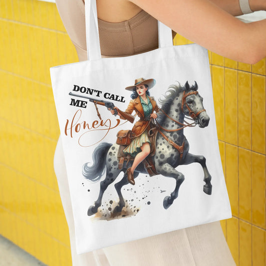 White tote bag with a fierce cowgirl on a horse, holding a gun, and bold text: “Don’t Call Me Honey.”