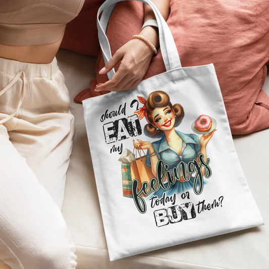 White tote bag with a retro pin-up girl holding a donut and shopping bags, with the quote:
“Should I eat my feelings today or buy them?”